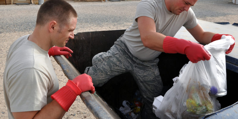 Two men in fatigues, one standing in the garbage bin