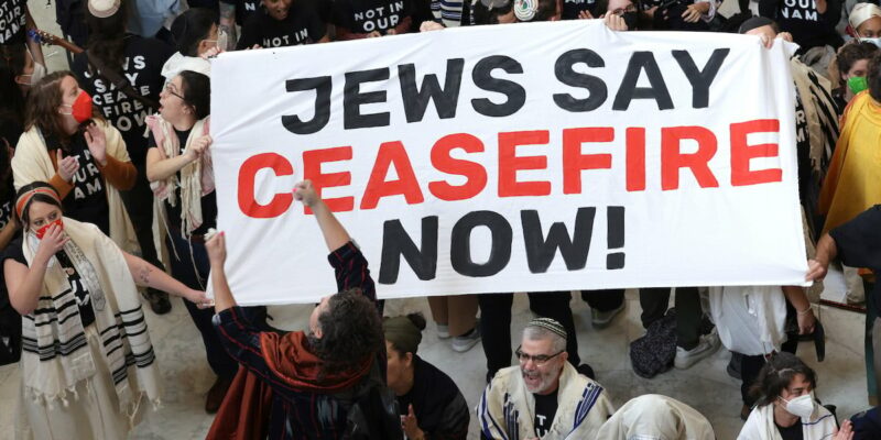 A crowd of protesters photographed from above. Some are wearing Jewish prayer shawls and yarmulkes. IN the middle is a sign with large letters reading "JEWS SAY CEASEFIRE NOW!"