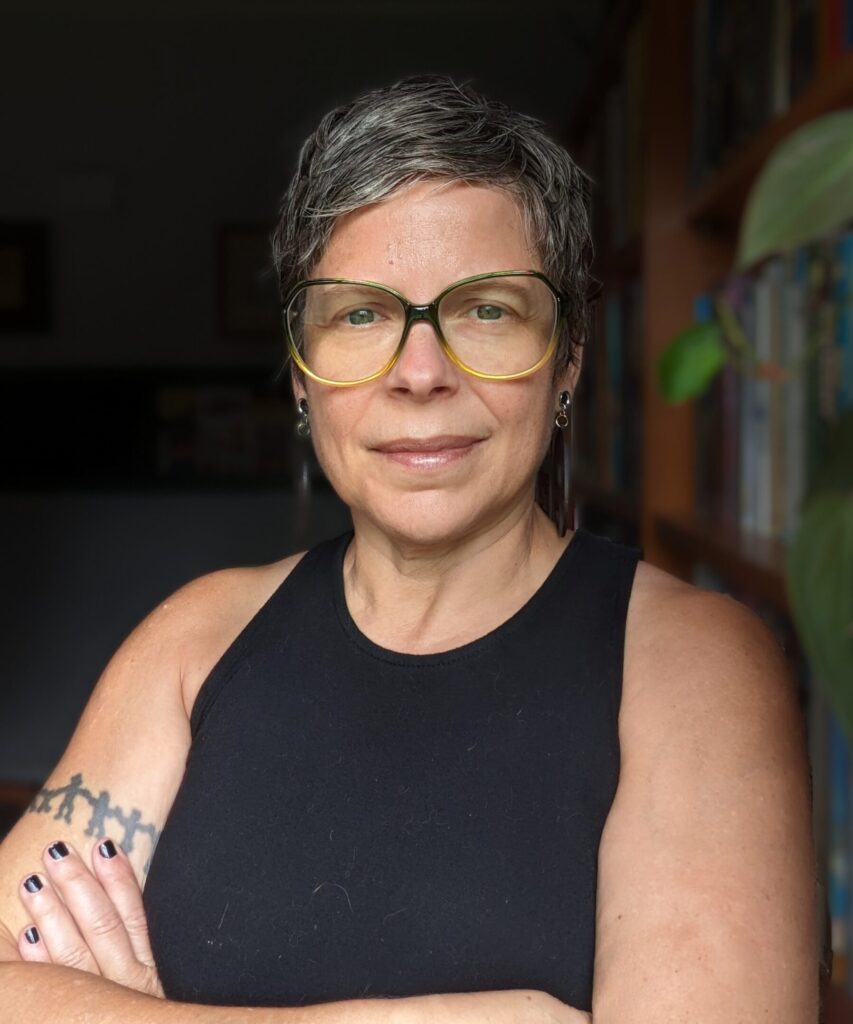 A white person with short gray-brown hair and large glasses stands in front of a bookcase with her arms crossed and a knowing smile on her face